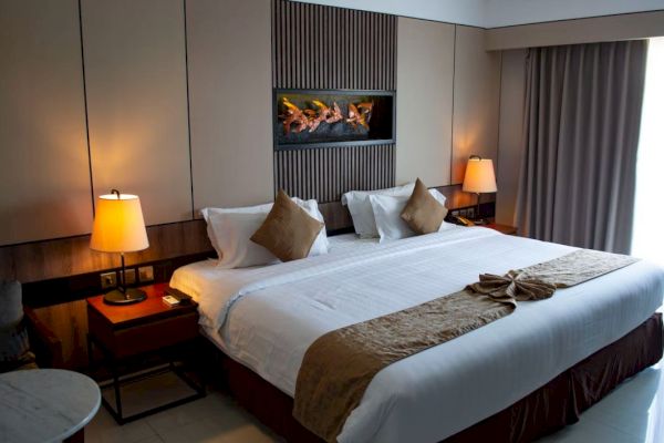 A modern hotel room with a large bed, decorative pillows, and warm lighting. A faux fireplace adds ambiance above the bed.