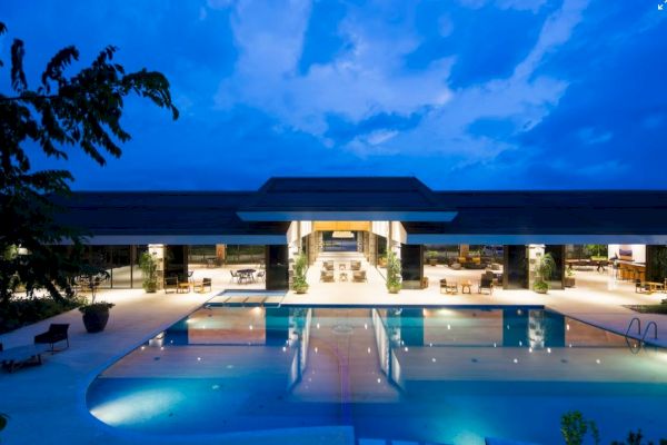 A modern building with a large, illuminated swimming pool at night, surrounded by lounge chairs and open spaces.