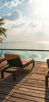 Wooden deck with loungers and umbrellas by a pool overlooking the ocean, surrounded by palm trees.
