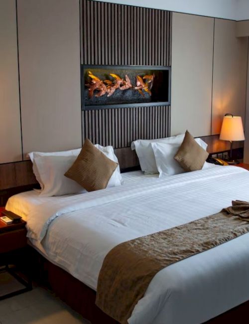 A modern hotel room with a large bed, decorative pillows, lamps on side tables, and a fireplace feature on the wall.