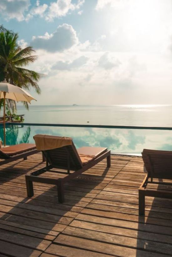 A tranquil poolside scene with sun loungers, umbrellas, palm trees, and a view of the ocean under a partly cloudy sky.