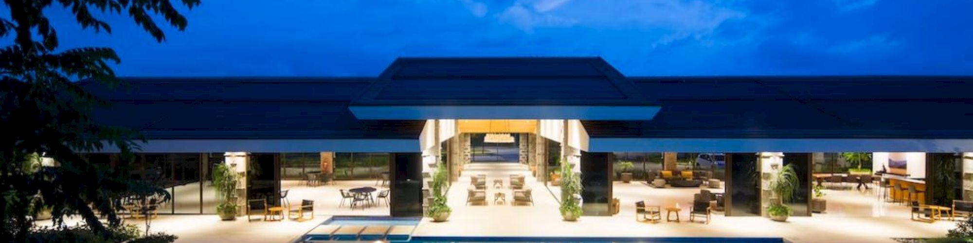 The image shows a luxurious, illuminated pool area with a modern building in the background under a blue evening sky.