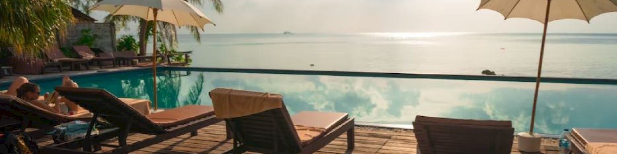 Lounge chairs and umbrellas by a serene infinity pool with a tropical backdrop and bright sky above.