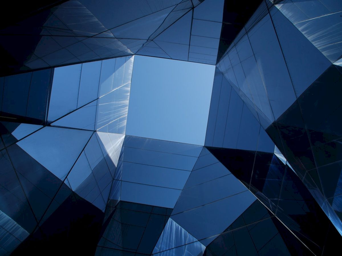 The image shows a geometric architectural structure with reflective glass surfaces, forming a skylight opening to the blue sky.