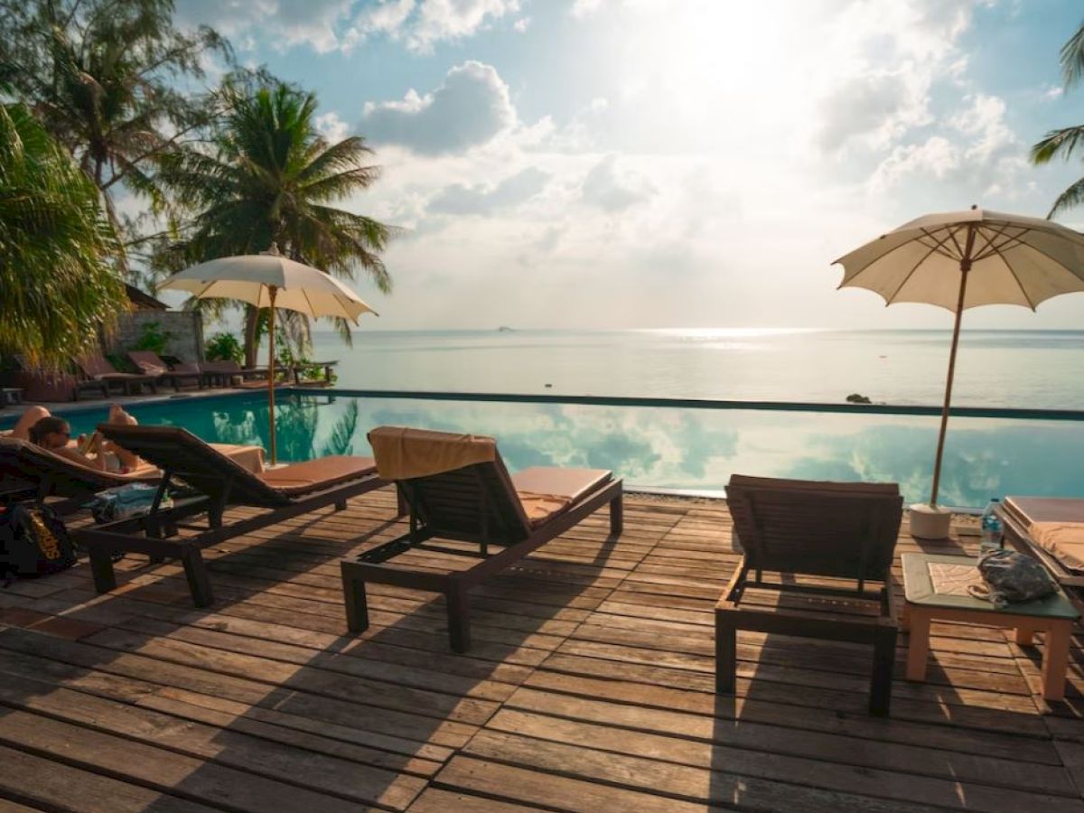 Poolside scene with lounge chairs and umbrellas overlooking a calm sea, with palm trees and a bright sky in the background.