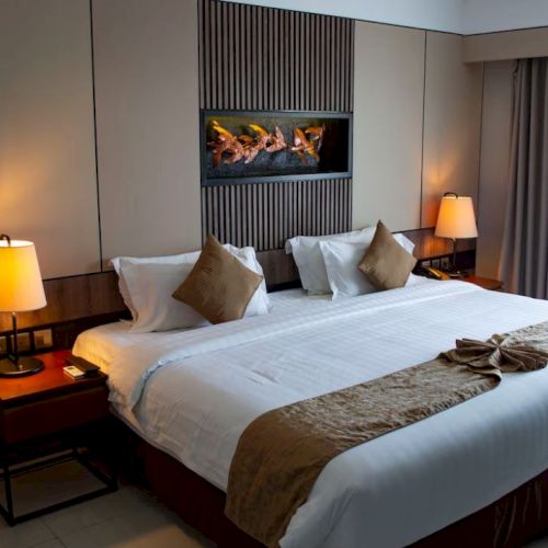 The image shows a neatly arranged hotel bedroom with a large bed, two lamps, a small table, and curtained window, creating a cozy atmosphere.