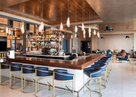 The image shows a modern bar with a wooden counter, blue bar stools, pendant lights, and a well-stocked liquor shelf in an open, stylish space.