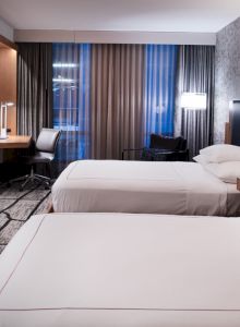 The image shows a modern hotel room with two beds, a TV, desk, and stylish lighting. The decor is contemporary with a patterned carpet.