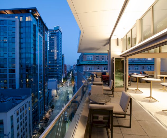 A modern rooftop patio with tables and chairs, overlooking a city street lined with tall buildings during twilight.