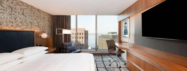 A modern hotel room with a large bed, desk, TV, and window offering a city view, featuring stylish decor and carpeted flooring.