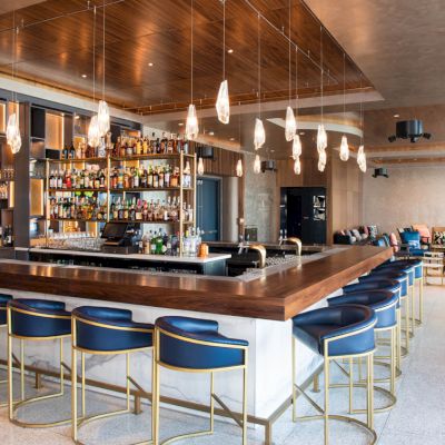 A modern bar with hanging lights, a wooden counter, blue bar stools, and a variety of bottles on shelves in the background.