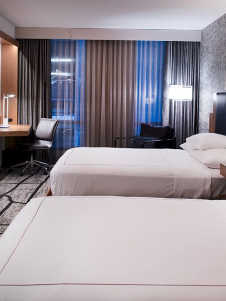 The image shows a modern hotel room with two beds, a flat-screen TV, a desk, and large windows with curtains.