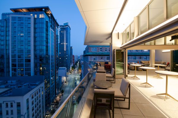 A modern city high-rise balcony at twilight, overlooking a street with tall buildings, featuring outdoor seating and tables.