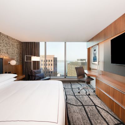 A modern hotel room with a large bed, big window, desk with chair, and wall-mounted TV, featuring a cityscape view in the background.