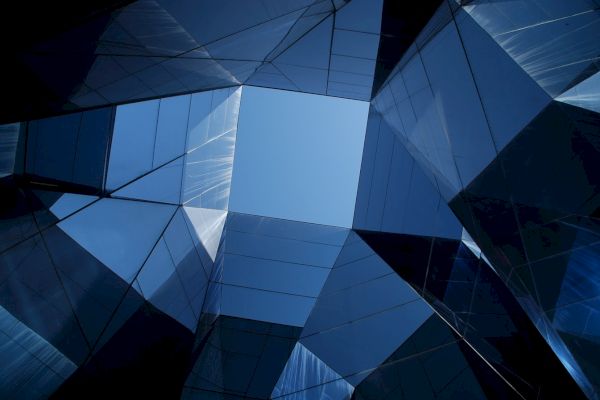 The image shows an architectural view upwards through dark blue glass panels forming geometric shapes with a central opening to the sky.