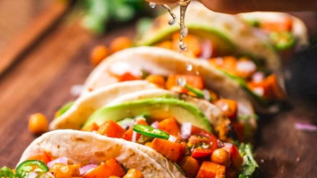 The image shows tacos filled with veggies and chickpeas, garnished with avocado slices and cilantro, with lime being squeezed on top.