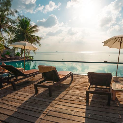 A serene beach scene with lounge chairs, umbrellas, palm trees, and an infinity pool overlooking a calm ocean and cloudy sky.