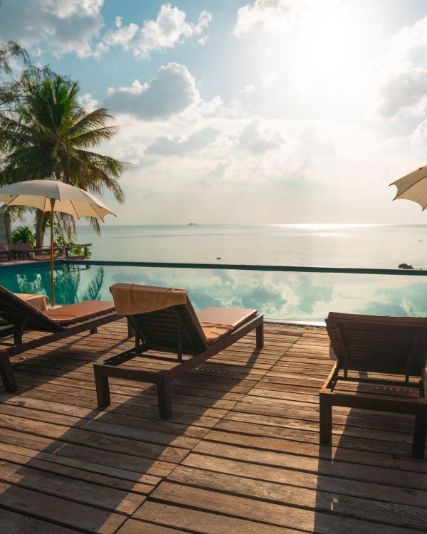 A serene beach scene with lounge chairs, umbrellas, palm trees, and an infinity pool overlooking a calm ocean and cloudy sky.