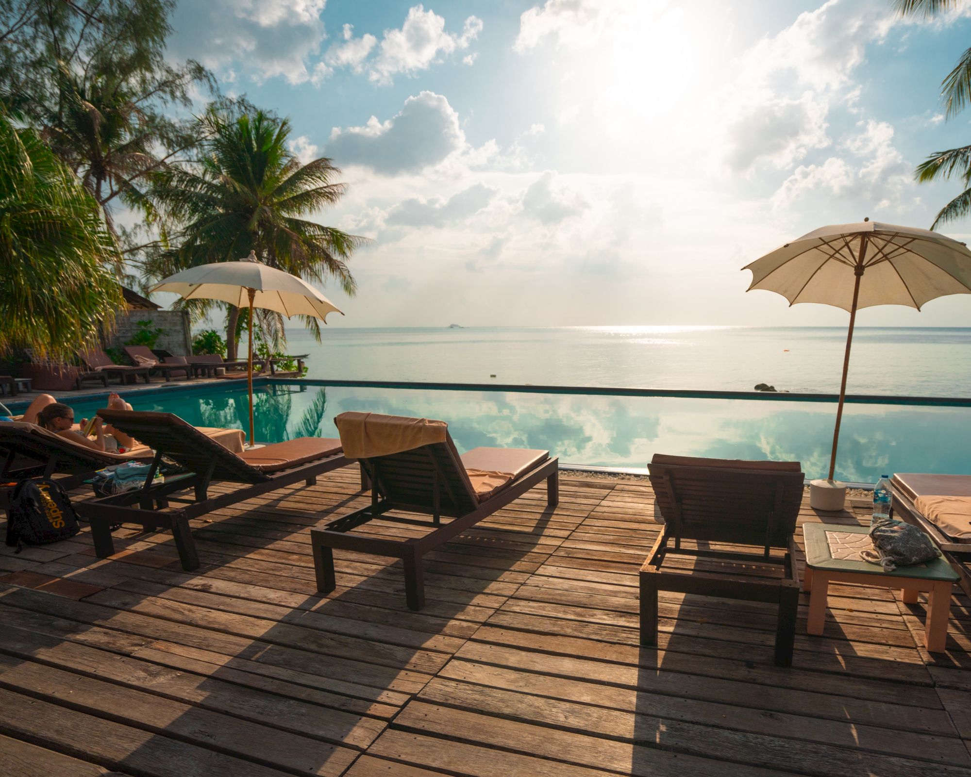 Lounge chairs and umbrellas line a wooden deck by an infinity pool, with a scenic view of the ocean and cloudy sky as the sun shines.
