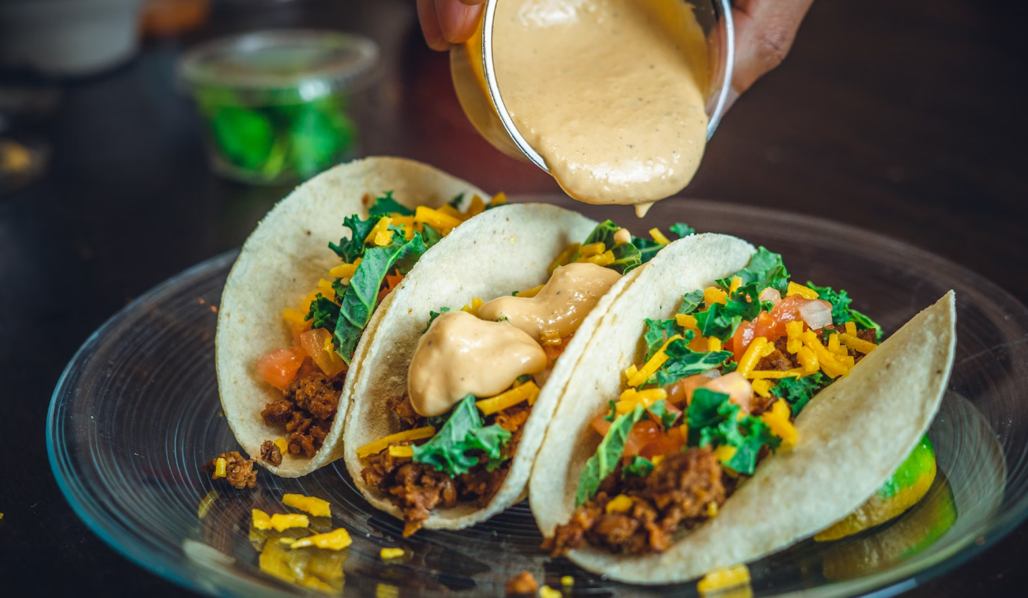 Three tacos filled with ground meat, greens, cheese, and tomatoes, topped with a creamy sauce on a clear plate.