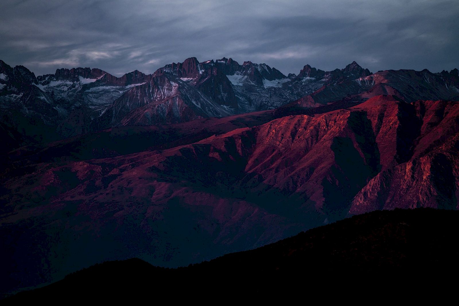 Mountain range with dramatic peaks, highlighted by red and purple hues under a cloudy sky at sunset or sunrise.