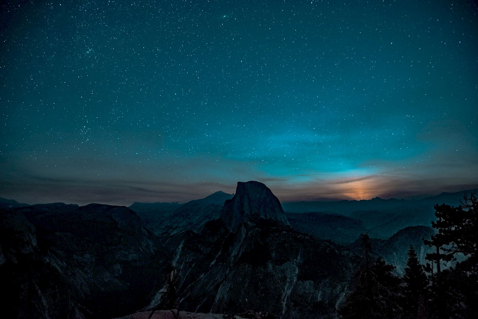 A starry night sky with visible stars is above a mountain landscape, featuring a prominent peak and silhouetted trees in the foreground.
