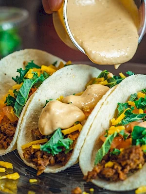 Three tacos with meat, cheese, lettuce, and tomato, topped with sauce being poured from a cup, all served on a plate.