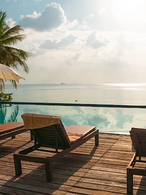 A serene beachside scene with lounge chairs, umbrellas, palm trees, and a calm ocean view under a partly cloudy sky.
