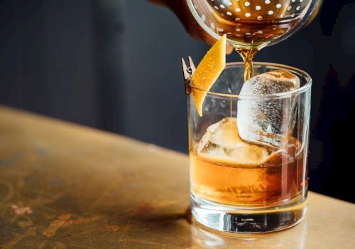 A cocktail being poured into a glass with ice and a lemon twist garnish on the rim, set on a wooden surface.