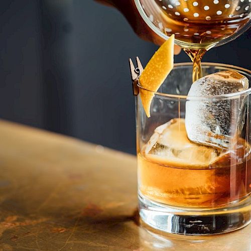 A cocktail being poured into a glass with ice and a lemon twist garnish on the rim, set on a wooden surface.