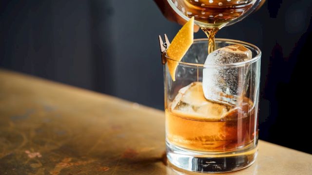 A cocktail being poured into a glass with ice and a lemon twist garnish on the rim, set on a wooden surface.