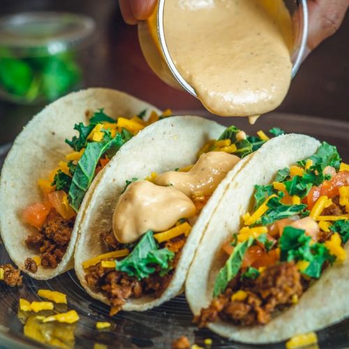 Three tacos with seasoned meat, cheese, lettuce, and chopped tomatoes being topped with a creamy sauce on a plate.