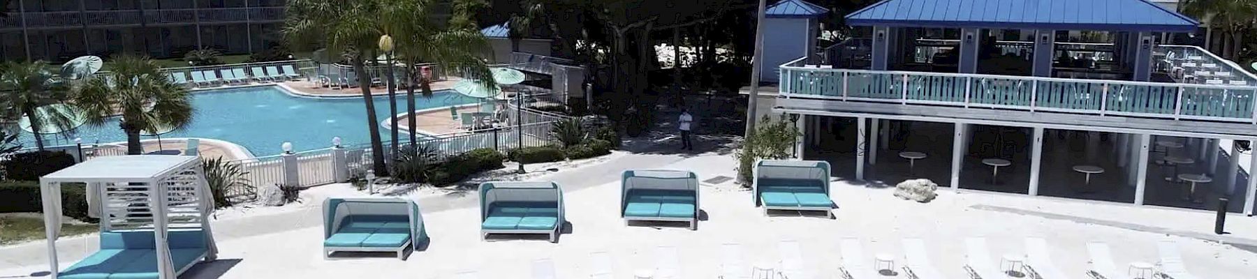 A beach resort scene with lounge chairs on sand, cabanas, a pool surrounded by palm trees, and a blue-roofed sky bar.