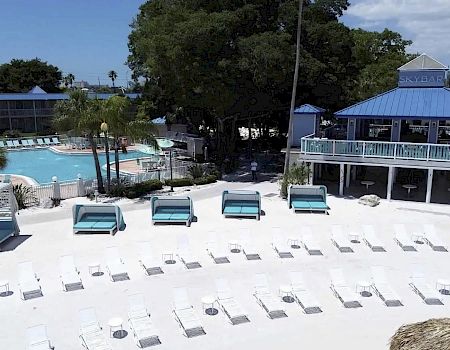 A beach resort scene with lounge chairs on sand, cabanas, a pool surrounded by palm trees, and a blue-roofed sky bar.