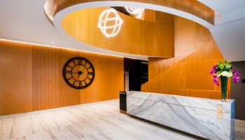 A modern interior with a marble desk, spiral wooden structure, large wall clock, and decorative lighting, featuring a flower arrangement.