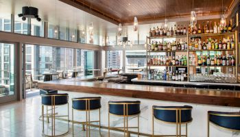 A modern bar with blue stools and a stocked liquor shelf, surrounded by windows providing a city view.