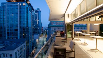 A cityscape at dusk, viewed from a modern balcony with chairs and tables, overlooking a street lined with tall buildings.