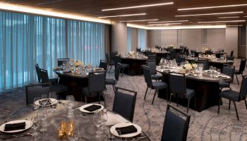 The image shows a banquet hall set with round tables, chairs, glassware, and floral arrangements, under modern lighting.