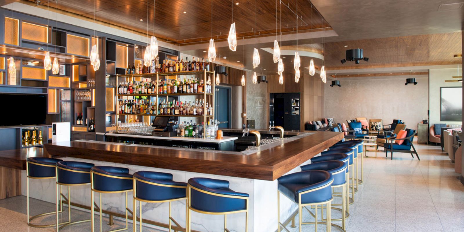 The image shows a modern bar with a wooden counter, blue barstools, pendant lights, and a well-stocked liquor shelf in a stylish lounge area.