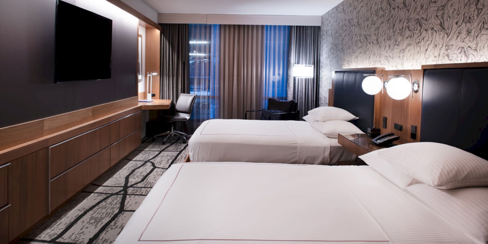 The image shows a modern hotel room with two double beds, a TV, a desk with a chair, and patterned wallpaper, creating a cozy atmosphere.