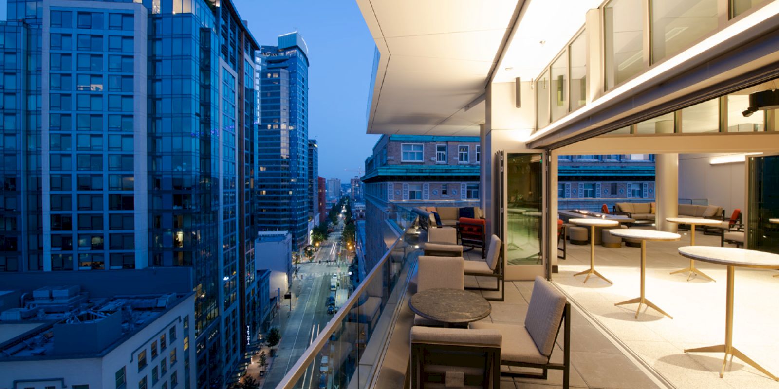 A modern rooftop terrace at dusk, overlooking a cityscape with tall buildings, featuring seating and tables along a glass railing.