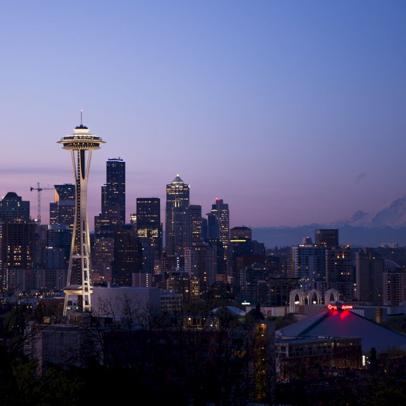 Seattle skyline at dusk with the Space Needle and Mount Rainier in the background, city lights illuminating the urban landscape.