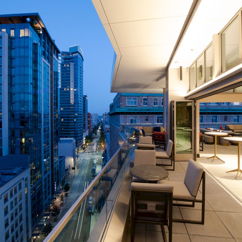 A modern rooftop balcony with seating overlooks a city street lined with tall buildings at dusk, featuring glowing interior lights.