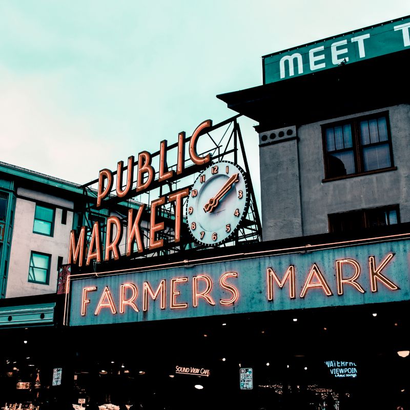 The image shows a building with neon signs for "Public Market" and "Farmers Market," with a clock in between, under a cloudy sky.