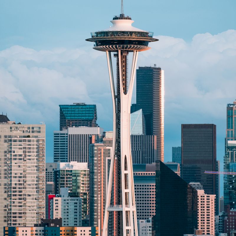 The image shows the Space Needle with a backdrop of urban buildings under a cloudy sky, showcasing a cityscape.