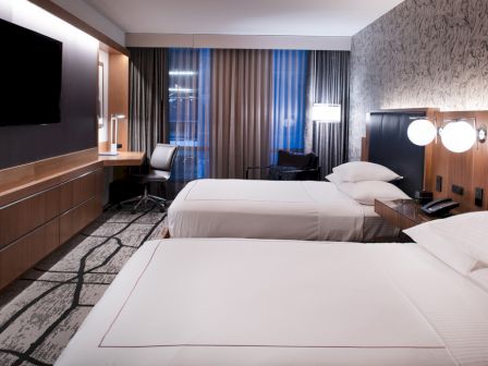 A modern hotel room with two double beds, a desk with a chair, a wall-mounted TV, and lamps, featuring stylish decor and carpeting.
