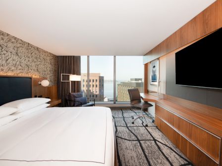 A modern hotel room with a bed, desk, chair, large TV, and a window view of buildings and water.