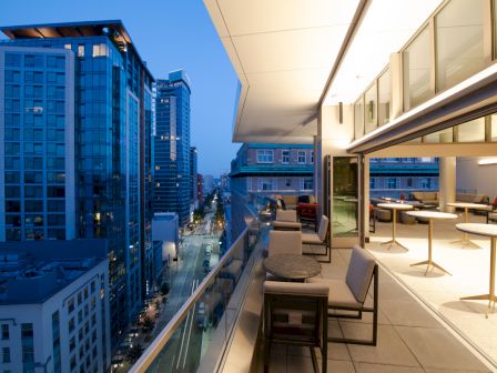 A rooftop terrace with seating overlooks a modern cityscape at dusk, featuring tall buildings and city lights along the street.