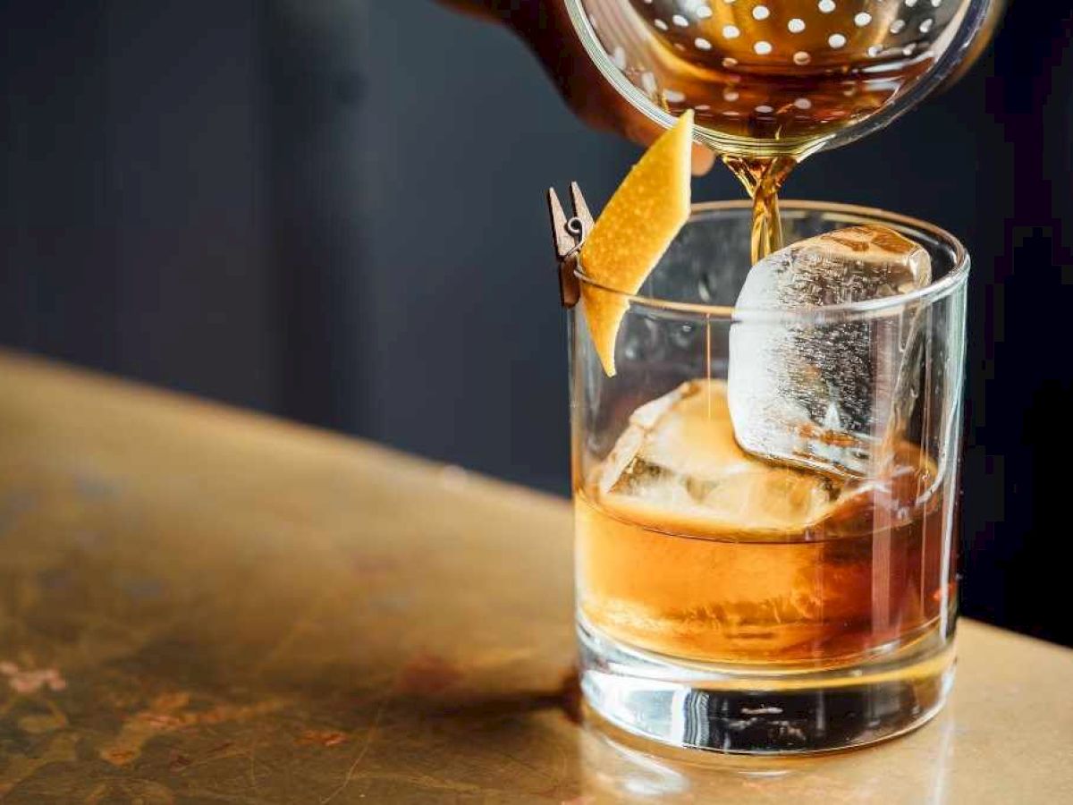 A cocktail is being poured into a glass with ice and garnished with a twist of citrus peel.
