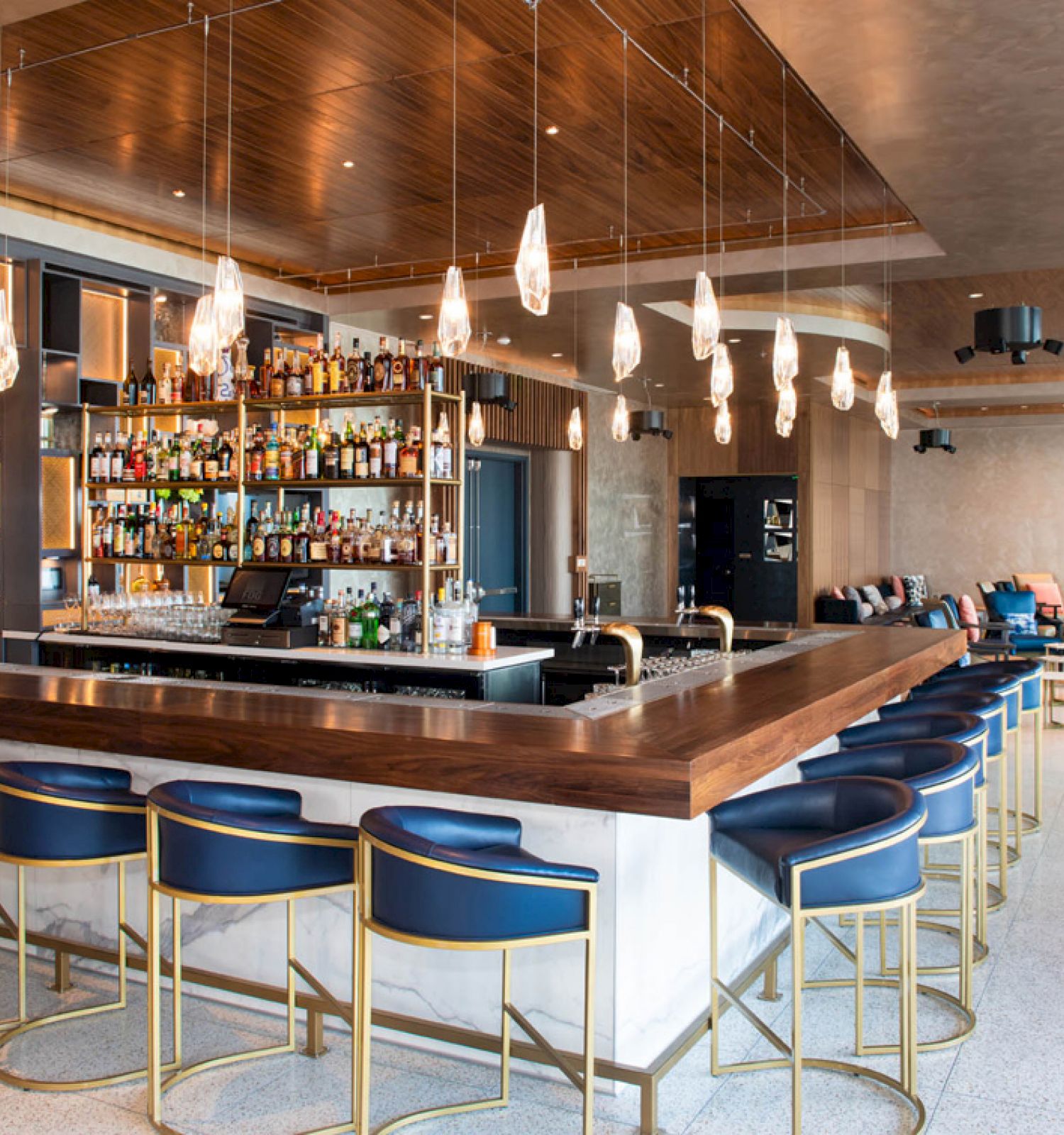 The image shows a modern bar with a wooden counter, blue barstools, and pendant lights. Shelves filled with bottles are behind the bar.